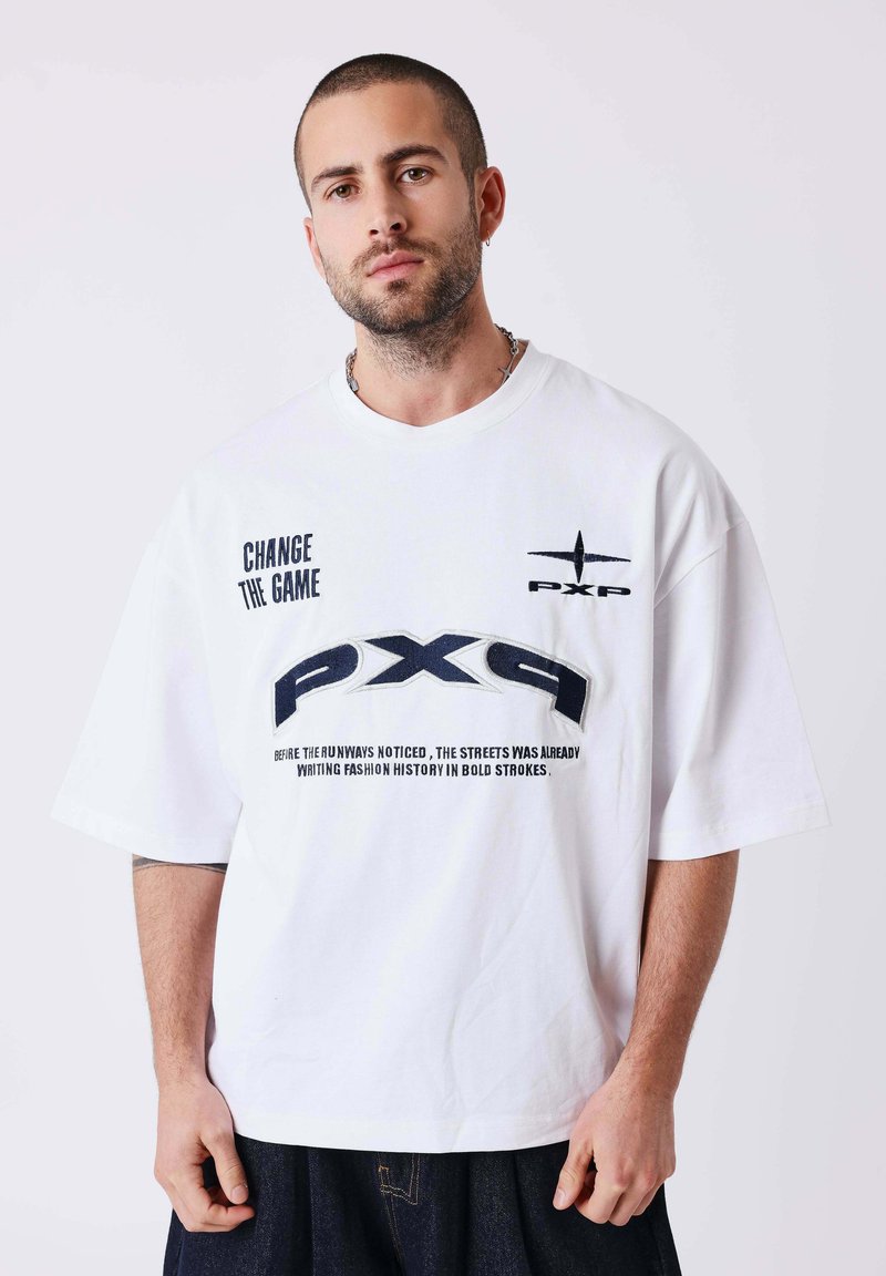Homme aux cheveux courts et barbe porte une chemise blanche oversize avec du texte noir "CHANGE THE GAME" et des logos "PXP" sur un fond uni.