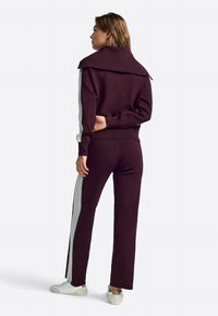 Bordo trenirka z visokim ovratnikom in svetlo modrimi črtastimi poudarki na rokavih in stranskih šivih, obuta s svetlimi čevlji.