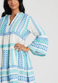 Vestido de manga larga en tela blanca con patrones geométricos azules. Presenta un cuello y una parte inferior con pliegues para añadir textura.