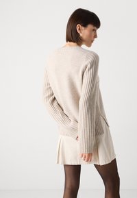 Lys beige genser med ribbet ermer, sidesliss, og en avslappet passform, sammen med en kremfarget plissert skjørt og sorte strømpebukser.