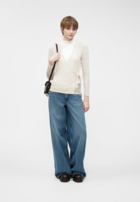 Beige wrap-toppi, jossa on pitkät hihat, yhdistettyinä suureisiin sinisiin farkkuihin. Malli pitää kädessään pientä mustaa olkalaukkua. Sileä kangas.