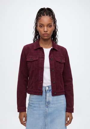 Jeune femme aux cheveux tressés portant une veste en velours côtelé bordeaux, une chemise blanche et une jupe en denim bleu clair, debout devant un fond blanc.