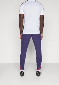 Sportliches Outfit mit einem weißen Kurzarmshirt mit schwarzen Akzenten und lila schmalen Hosen mit pinken Seitenstreifen, von hinten angezeigt.