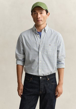 Mann mit grünem Cap, weiß-blau kariertem Hemd mit hochgekrempelten Ärmeln und dunkelblauer Jeans, stehend vor neutralem Hintergrund.