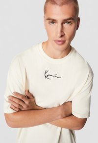 Kratka krem t-shirt s kratkim rukavima od pamuka, s crnim izvezenim logotipom potpisom na prsima, s opuštenim krojem.