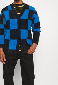 Huppari, jossa on siniharmaa ruudullinen kuvio, V-kaula-aukko ja mustat napit, päällä musta ja keltainen raidallinen paita.