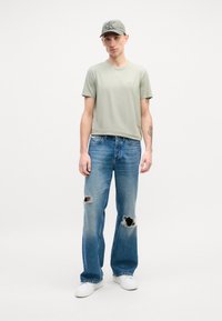 T-shirt vert clair, coupe décontractée ; jean bleu délavé avec de grands trous ; baskets blanches ; casquette grise avec logo ; fond minimaliste.