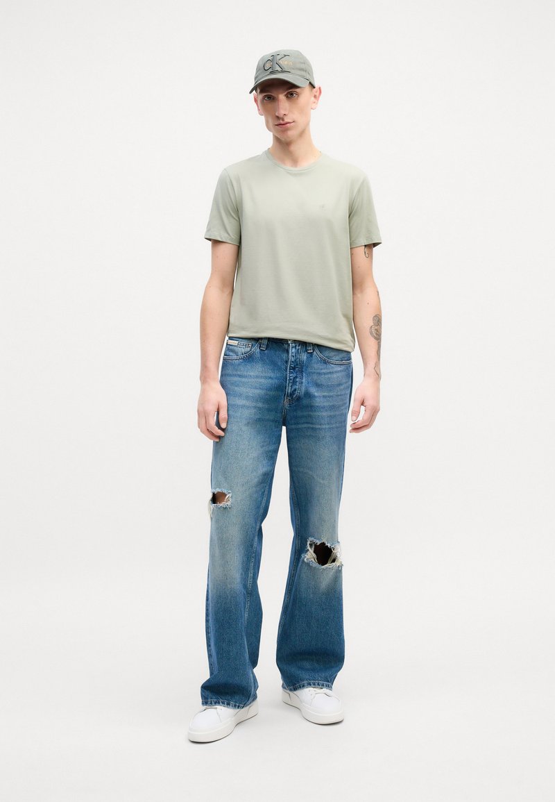 Light green T-shirt, relaxed fit; faded blue jeans with large holes; white sneakers; gray cap with logo; minimalist background.