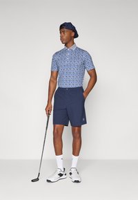 Poloko s krátkým rukávem s modrým vzorem v kombinaci s námořnickými šortkami. Model drží golfovou hůl, má na sobě bílé a černé sportovní boty a čepici.