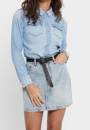 Kobieta w jasnoniebieskiej koszuli jeansowej na guziki, włożonej w wyblakłą spódnicę jeansową z czarnym paskiem, jedna ręka w kieszeni, druga oparta na spódnicy.