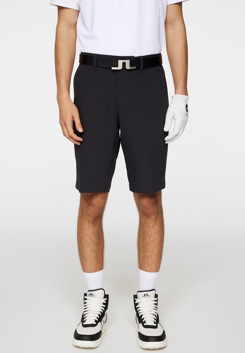 Svarta golfshorts med en slät textur, resårmidja med en silverfärgad spänne, tillsammans med en vit skjorta och svarta högklackade skor.