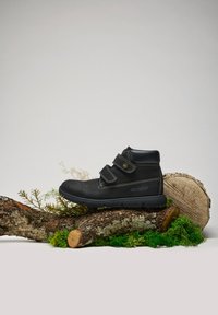 Bottines noires en cuir au niveau de la cheville avec deux sangles ajustables, surpiqûres blanches contrastantes, surface texturée et semelle en caoutchouc, posées sur une bûche.