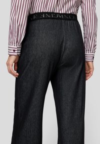 Mann trägt dunkle Nadelstreifenhose mit schwarzem elastischem Bund und lila-weiß gestreiftes Hemd mit vertikalen Streifen und geknöpften Manschetten.