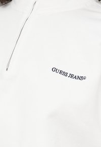 Top in cotone bianco con zip, dotato di una zip a un quarto di lunghezza e un logo ricamato in nero "GUESS JEANS" sul lato sinistro.