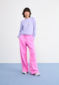 Sweat-shirt lilas avec logo Nike, associé à un pantalon de survêtement rose à coupe ample. L'ensemble est complété par des baskets grises et blanches sur un fond bleu.