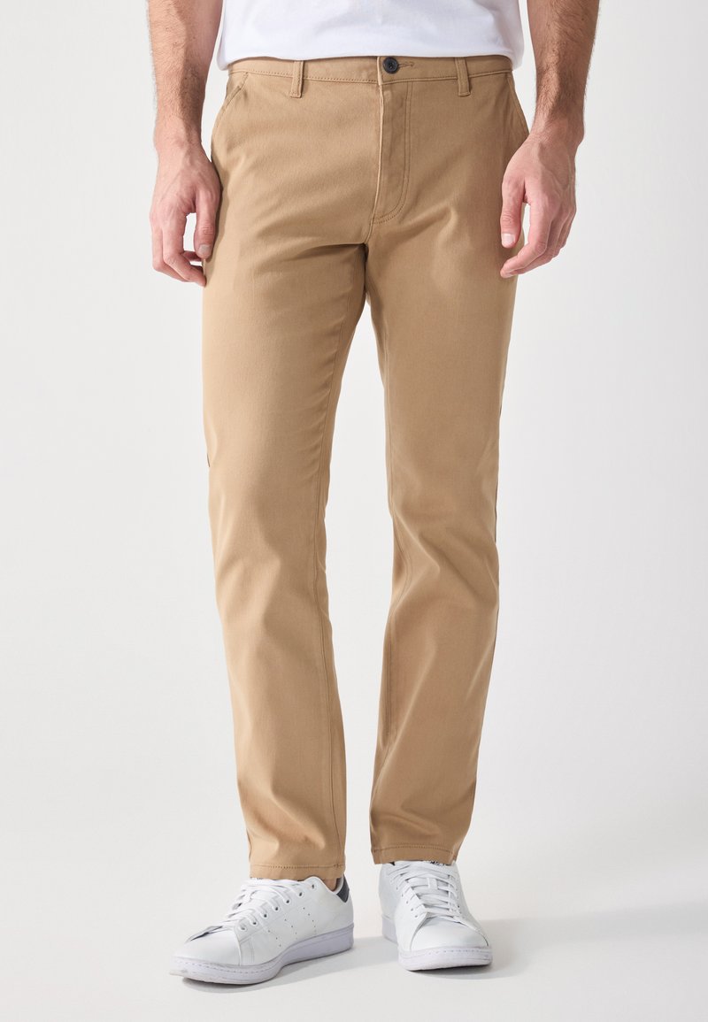 Chino in cotone beige con vestibilità dritta, dotati di una chiusura anteriore con un singolo bottone e tasche laterali. Indossati con sneakers bianche.
