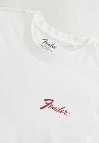 Fehér póló piros Fender logóval az elején, valamint fekete Fender logóval és védjeggyel a gallér belsejében.