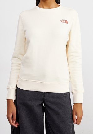 Woman wearing a cream long-sleeve sweatshirt with a small red "The North Face" logo on the left chest and dark gray pants.