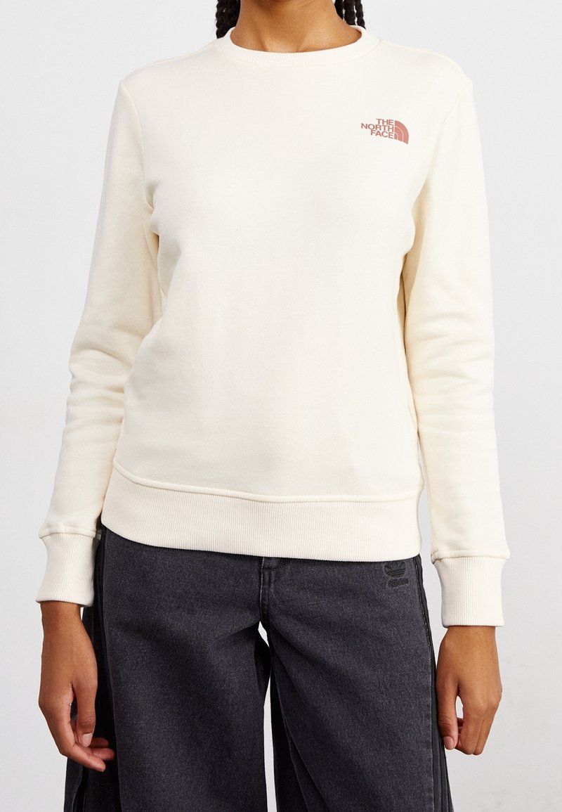 Femme portant un sweat-shirt à manches longues crème avec un petit logo rouge "The North Face" sur la poitrine gauche et un pantalon gris foncé.