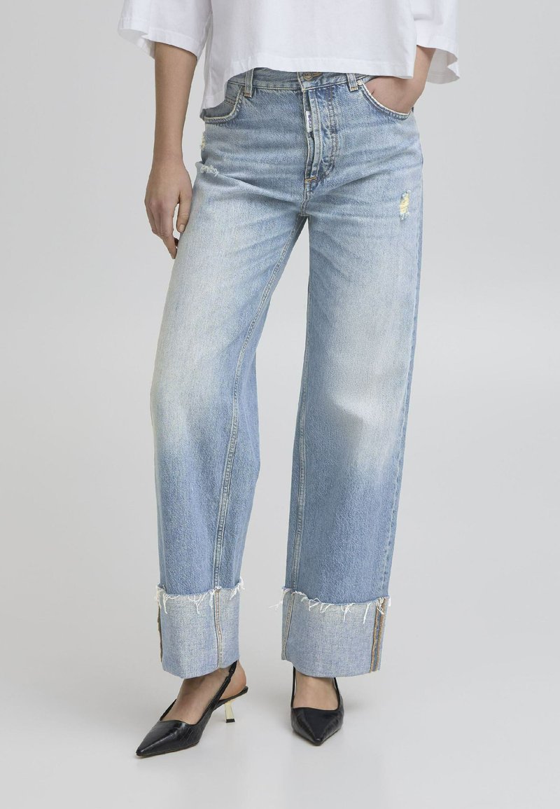 Helle blaue Jeans mit weitem Bein, fransigem Saum, hoher Taille und subtilen Abriebstellen. Kombiniert mit schwarzen, spitz zulaufenden Pumps.