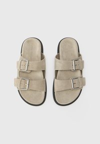 Două sandale din suede bej, cu talpă plată, având două curele late cu catarame argintii ajustabile și o talpă din cauciuc texturat negru.