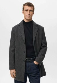 Manteau gris texturé avec un col cranté et trois boutons, porté sur un col roulé noir, associé à un jean foncé et une ceinture noire.