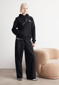 Felpa nera con tasca frontale e logo verde, abbinata a pantaloni larghi neri e scarpe a fantasia bianco e nero, su uno sfondo neutro.