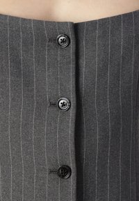 Šedá pruhovaná tkanina má čtyři černé knoflíky vpředu, přičemž jemné pruhy dodávají texturu a detail tomuto oděvu.