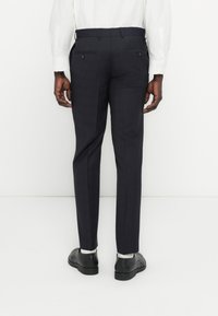 Tamsiai mėlynos, kostiuminės kelnės tiesaus kirpimo, su dviem kišenėmis gale ir lygiu audinio paviršiumi. Dėvimos su juodais batais ir baltomis kojinėmis.