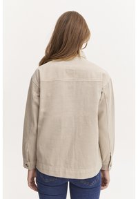 Beżowa bawełniana kurtka o luźnym kroju, z kołnierzem, zapinanymi na guziki mankietami i gładką fakturą, noszona na niebieskie jeansy o prostym wzorze.