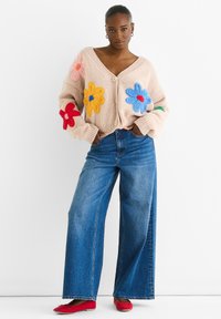 Beige knitted cardigan with colorful floral appliqués, paired with wide-leg blue denim jeans and red flat shoes. Button closure at front.