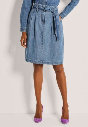 Vrouw met een blauwe spijkerrok tot aan de knie met een gestrikte riem, bijpassend denimshirt en fel paarse hoge hakken, staand tegen een effen achtergrond.