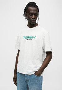 Ανοιχτό γκρι βαμβακερό μπλουζάκι με κοντά μανίκια, με έντονο γραφικό "TOMMY JEANS" σε τιρκουάζ και μαύρο. Χαλαρή εφαρμογή και λαιμόκοψη τύπου crew.