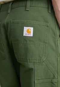 Groene katoenen twill broek met een achterzak voorzien van een wit vierkant Carhartt logo patch. De stof heeft een stevige, gestructureerde uitstraling.