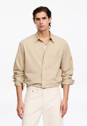 Jeune homme aux cheveux foncés portant une chemise beige à manches longues boutonnée avec les manches retroussées et un pantalon de couleur claire sur fond blanc.