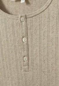 Vakrameszövetből készült bézs kötött póló, kerek nyakkivágással, függőleges csíkokkal és gyémántmintával, három fehér gombbal rögzítve.