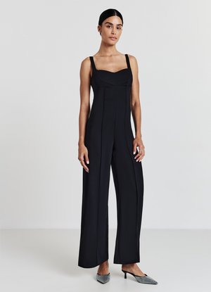 Schwarzer Jumpsuit mit quadratischem Ausschnitt, weitem Bein und Nahtdetails. Kombiniert mit spitz zulaufenden Schuhen für einen eleganten Look.