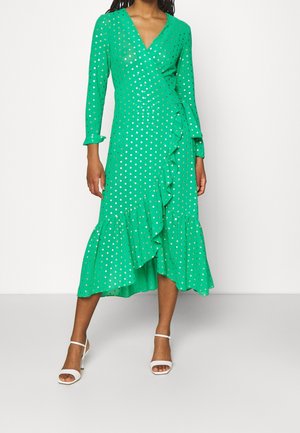 Femme portant une robe portefeuille verte à pois dorés, ourlet à volants et sandales blanches à talons, debout sur un fond blanc uni.