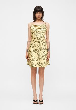 Γυναίκα με μεσαίου μήκους σκουρόχρωμα μαλλιά, φορώντας ένα κίτρινο αμάνικο φόρεμα με μαύρες λεοπάρ κηλίδες και μαύρες ανοιχτές γόβες, στέκεται μπροστά σε λευκό φόντο.