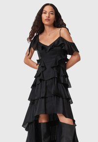 Donna con lunghi capelli ricci che indossa un vestito nero a maniche scoperte a strati e stivali neri al ginocchio su uno sfondo neutro.