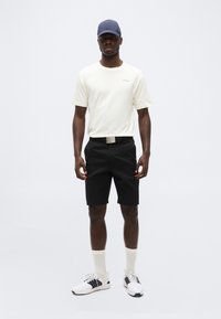 Άνδρας στέκεται φορώντας λευκό πουκάμισο με κοντά μανίκια, μαύρη σορτς με ζώνη, λευκές κάλτσες, λευκά αθλητικά παπούτσια και μπλε καπέλο μπέιζμπολ, μπροστά σε λευκό φόντο.