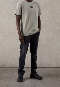 Camiseta de algodón verde claro con un parche de logo rectangular en rojo, blanco y azul. Combinada con jeans gris oscuro y zapatillas negras.