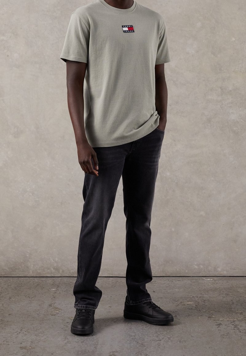 Camiseta de algodón verde claro con un parche de logo rectangular en rojo, blanco y azul. Combinada con jeans gris oscuro y zapatillas negras.