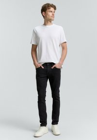 Camiseta blanca de manga corta combinada con jeans negros ajustados. El modelo lleva zapatillas blancas, con las manos en los bolsillos, de pie contra un fondo gris claro.