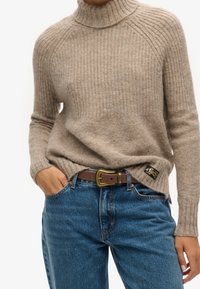 Person wearing beige ribbed turtleneck sweater tucked into blue jeans with brown leather belt and hand in pocket.