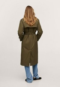 Oliven grøn trenchcoat med bælte om taljen, dobbeltknappet design, skulderklapper og en rygventilation. Matchet med blå flagrende jeans.