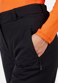 Pantaloni neri in materiale elastico con tasca con zip, dotati di vita piatta e design aderente, abbinati a un top arancione a maniche lunghe.