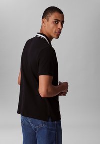 Black polo shirt with a white striped collar, short sleeves, and a straight hem. Worn with blue jeans, showcasing a casual design.