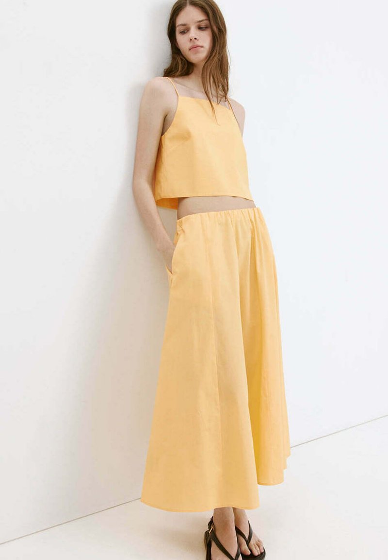 Jeune femme portant un top court jaune et une jupe large, debout les mains dans les poches contre un mur blanc.