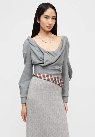 Γκρι φούτερ με ντραπαρισμένο λαιμόκοψη και μακριά μανίκια, σε συνδυασμό με υφασμάτινη γκρι φούστα με υφή. Κόκκινο και πράσινο καρό διακοσμητικό γύρω από τη μέση.